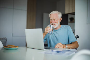 Senior man in a wheelchair working from home during retirement. Elderly man using digital technologies, working on a laptop, videocalling someone. Concept of seniors and digital skills. - HPIF34204