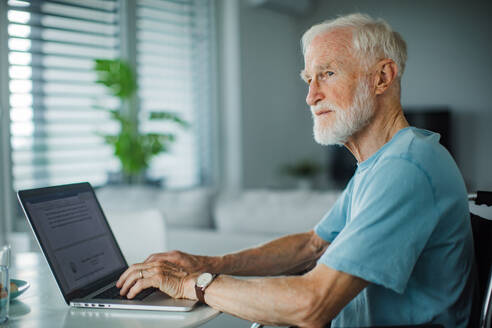 Senior man in a wheelchair working from home during retirement. Portrait of elderly man using digital technologies, working on a laptop. Concept of seniors and digital skills. - HPIF34208