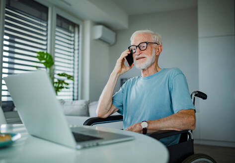 Senior man in a wheelchair working from home during retirement. Elderly man using digital technologies, working on a laptop and making phone call at home. Concept of seniors and digital skills. - HPIF34210