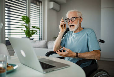 Älterer Mann im Rollstuhl, der im Ruhestand von zu Hause aus arbeitet. Älterer Mann, der digitale Technologien nutzt, an einem Laptop arbeitet und zu Hause telefoniert. Konzept von Senioren und digitalen Fähigkeiten. Senior, der sich beim Kundendienst beschwert. - HPIF34211