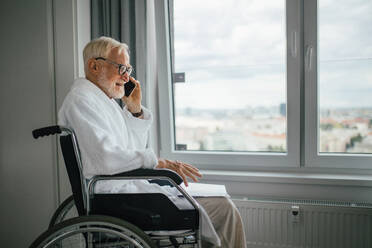 Älterer Mann im Rollstuhl, der morgens im Bademantel telefoniert. Konzept der Einsamkeit und Abhängigkeit von Menschen im Ruhestand. - HPIF34217