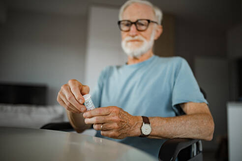 Ein älterer Mann im Rollstuhl nimmt zu Hause Tabletten ein. Ein chronisch kranker Mann nimmt Medikamente ein und liest das Etikett auf der Flasche. - HPIF34221