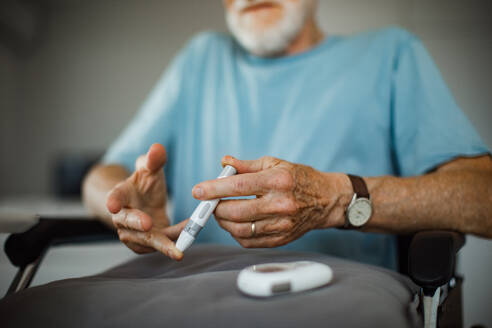 Diabetic senior man on wheelchair checking his blood sugar level with fingerstick urbaning glucose meter. Portrait of elderly man with type 1 diabetes using blood glucose monitor at home. - HPIF34223