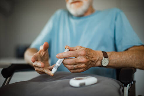 Diabetischer älterer Mann im Rollstuhl, der seinen Blutzuckerspiegel mit einem Glukosemessgerät mit Fingerstich kontrolliert. Porträt eines älteren Mannes mit Typ-1-Diabetes, der zu Hause ein Blutzuckermessgerät benutzt. - HPIF34223