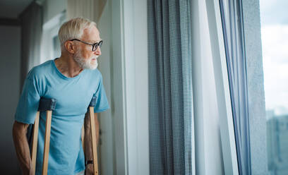 Sad senior man with crutches spending time alone in his apartment, looking out of window. Concept of loneliness and dependence of retired people. - HPIF34227