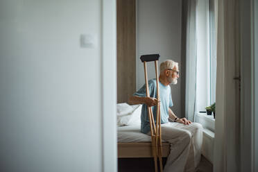 Sad senior man with crutches spending time alone in his apartment, looking out of window. Concept of loneliness and dependence of retired people. - HPIF34228