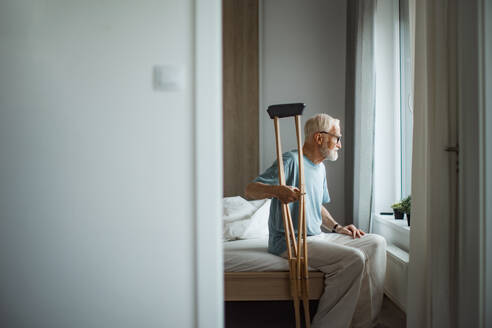 Sad senior man with crutches spending time alone in his apartment, looking out of window. Concept of loneliness and dependence of retired people. - HPIF34228