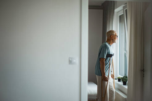Sad senior man with crutches spending time alone in his apartment, looking out of window. Concept of loneliness and dependence of retired people. - HPIF34229