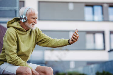 Senior man exercising outdoors in the city. Elderly man resting on a bench, listening to music through wireless headphones, taking selfie. - HPIF34238