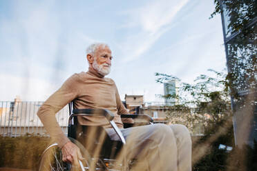 Älterer Mann im Rollstuhl sitzt draußen in einem städtischen Garten und genießt einen warmen Herbsttag. Porträt eines eleganten älteren Mannes mit grauen Haaren und Bart in einem Dachgarten in der Stadt. - HPIF34240