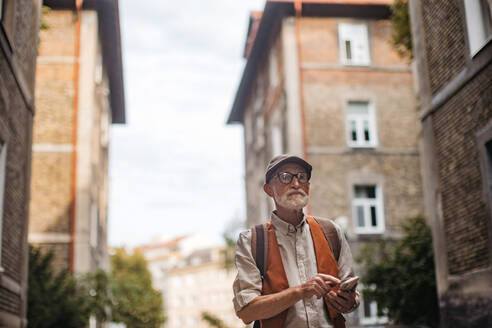 Älterer Tourist, der eine neue Stadt erkundet und interessante Sehenswürdigkeiten besucht. Älterer Mann, der mit einem Smartphone in der Hand durch die Stadt schlendert. - HPIF34249