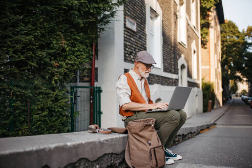 Senior man sitting on street curb working on his laptop outdoors. Portrait of elderly man using digital technologies, working on a notebook. Concept of seniors and digital skills. - HPIF34259