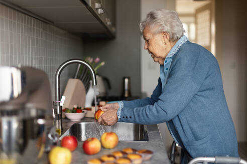 Glückliche ältere Frau, die in der Küche Äpfel wäscht, während die Großmutter Backwaren, Muffins, Kuchen und Süßigkeiten zubereitet. - HPIF34265