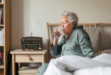 Senior woman sitting in pajama on a bed and drinkig water. - HPIF34286