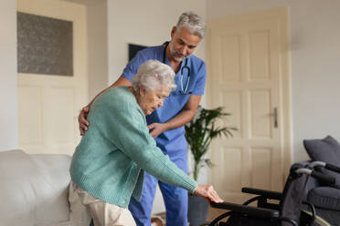 Eine ältere Frau versucht geduldig, aus ihrem Rollstuhl aufzustehen, und wird dabei von einer Pflegekraft unterstützt. - HPIF34309