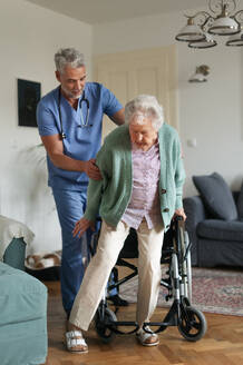 Caregiver helping senior woman to stand from her wheelchair in her home. An elderly woman is patiently trying to get up from her wheelchair. - HPIF34310