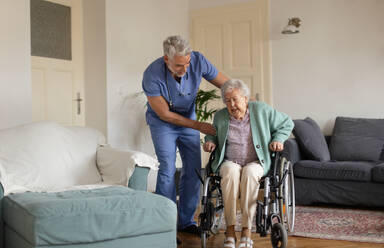 Caregiver helping senior woman to stand from her wheelchair in her home. An elderly woman is patiently trying to get up from her wheelchair. - HPIF34311