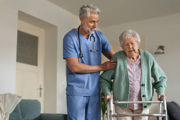 Caregiver helping senior woman to walk in her home. Thoughful male nurse taking care of eldery patient with walker. - HPIF34318