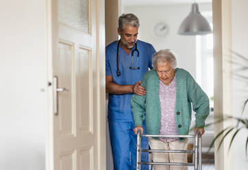 Ein Pfleger hilft einer älteren Frau beim Gehen in ihrem Haus. Ein umsichtiger Pfleger kümmert sich um einen älteren Patienten mit Rollator. - HPIF34320