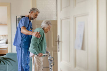 Ein Pfleger hilft einer älteren Frau beim Gehen in ihrem Haus. Ein umsichtiger Pfleger kümmert sich um einen älteren Patienten mit Rollator. - HPIF34321