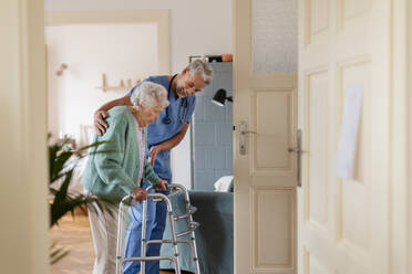 Ein Pfleger hilft einer älteren Frau beim Gehen in ihrem Haus. Ein umsichtiger Pfleger kümmert sich um einen älteren Patienten mit Rollator. - HPIF34322