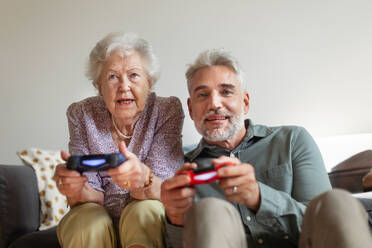 Mature man playing video games with his senior mother, having fun. Adult son enjoys spending time with his elderly mother. - HPIF34350