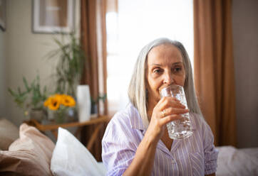 Portrait of senior woman sitting on the bed in a pajamas and drinking water. - HPIF34399