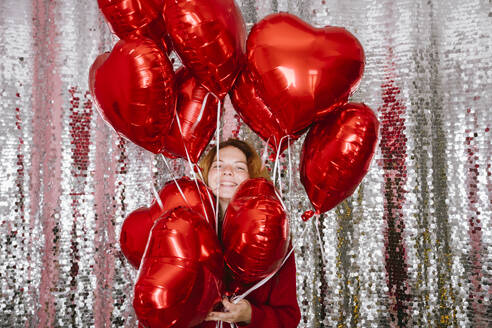 Happy woman holding heart shaped balloons in front of sequin curtain - YBF00326