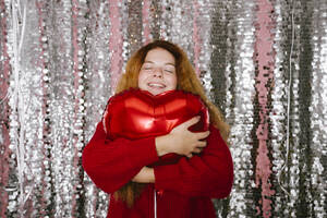 Happy woman hugging red balloon in front of sequin curtain - YBF00329