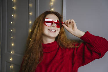 Smiling young woman wearing heart shaped sunglasses at home - YBF00331