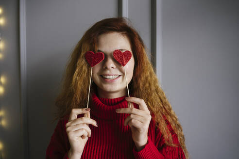 Playful woman holding heart shaped props over eyes at home - YBF00332