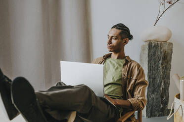 Thoughtful young man with laptop relaxing with feet on table at home - YTF01486