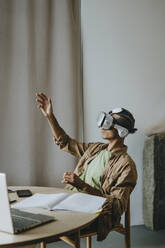 Junger Mann mit VR-Simulator sitzt mit erhobenem Arm am Tisch - YTF01501