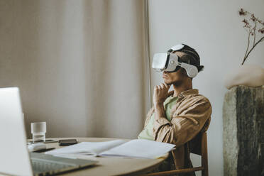 Young man wearing VR headset sitting with hand on chin at table - YTF01502