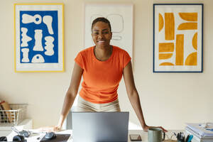 Lächelnde Geschäftsfrau lehnt sich an einen Schreibtisch mit Laptop in einem modernen Heimbüro - EBSF04230