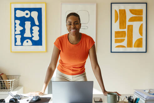 Smiling businesswoman leaning on desk with laptop in modern home office - EBSF04230