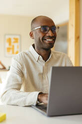 Lächelnder Geschäftsmann mit Brille, der einen Laptop am Schreibtisch in einem modernen Heimbüro benutzt - EBSF04241