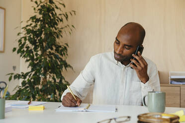 Geschäftsmann schreibt in Tagebuch und hört Smartphone im Büro zu Hause - EBSF04251