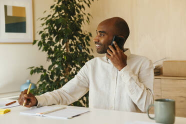 Smiling businessman listening to smart phone sitting with diary at home office - EBSF04252