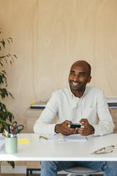 Smiling businessman sitting with smart phone at desk in home office - EBSF04253