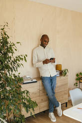 Lächelnder Geschäftsmann, der mit gekreuzten Beinen steht und ein Smartphone benutzt, das an der Wand im Heimbüro lehnt - EBSF04255