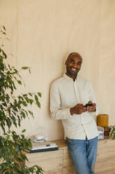 Lächelnder Geschäftsmann mit Smartphone an der Wand lehnend im Heimbüro - EBSF04256