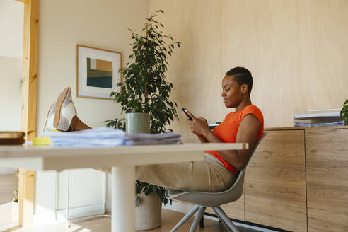 Relaxed businesswoman using smart phone with feet up on desk at home office - EBSF04259