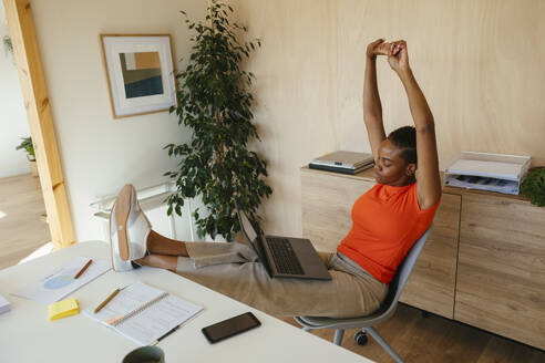Businesswoman with laptop stretching arms and feet up on desk at home office - EBSF04261