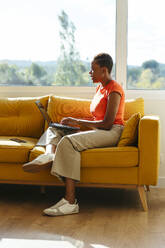 Businesswoman using laptop sitting on sofa by window at home office - EBSF04264