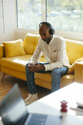 Geschäftsmann mit drahtlosen Kopfhörern und Smartphone auf dem Sofa im Büro zu Hause - EBSF04265