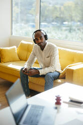 Businessman wearing wireless headphones sitting with smart phone on sofa at home office - EBSF04266