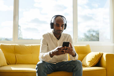 Geschäftsmann, der über drahtlose Kopfhörer mit seinem Smartphone auf dem Sofa im Büro zu Hause Musik hört - EBSF04268