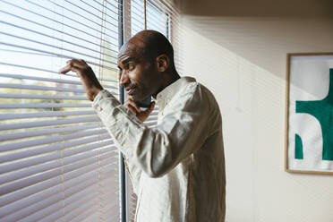 Mann schaut durch Jalousien aus dem Fenster und telefoniert zu Hause mit seinem Handy - EBSF04277
