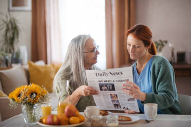 Krankenschwester verbringt Zeit mit einer älteren Frau zu Hause, liest gemeinsam Zeitung und bespricht die neuesten Nachrichten. Weibliche Pflegekraft kümmert sich um einen älteren Patienten. - HPIF34424